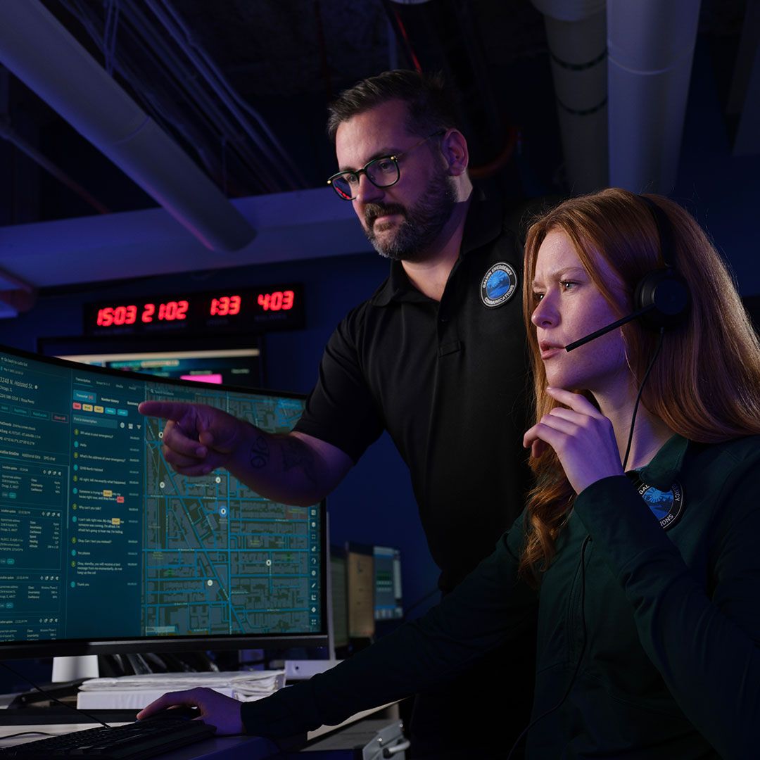 Two emergency dispatchers work together in a darkened control room. A man stands and points at a large computer monitor displaying a map and chat logs, while a woman wearing a headset sits at the station, looking intently at the screen.