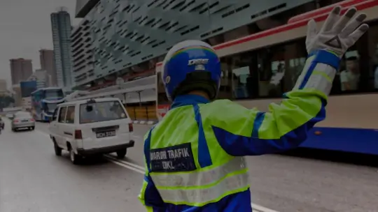 A person in a bright jacket and motorcycle helmet standing in a city street with their right arm raised.